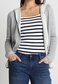 Kvinna klädd i en grå huvjacka över en svartvit randig ribbad ärmlös topp och blå denimjeans med händerna i fickorna.