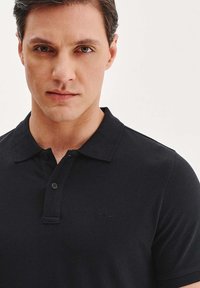 Czarna koszulka polo z klasycznym kołnierzykiem, zapięciem na dwa guziki i dyskretnym, strukturalnym materiałem. Małe logo na lewej piersi.