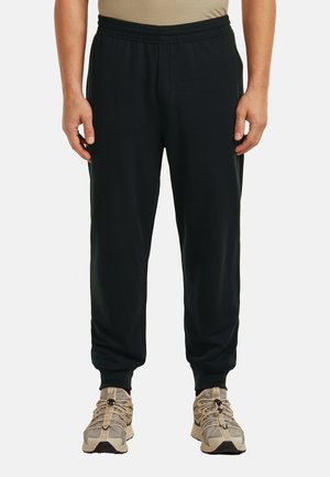 Mężczyzna w czarnych spodniach dresowych z elastycznymi mankietami, w beżowo-czarnych butach sportowych, stojący na jednolitym tle.