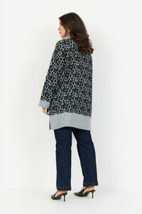 Frau mit langen braunen Haaren, die eine graue und schwarze Leopardenmuster-Cardigan, dunkle Jeans und schwarze High Heel Mules trägt, seitlich vor einem weißen Hintergrund stehend.