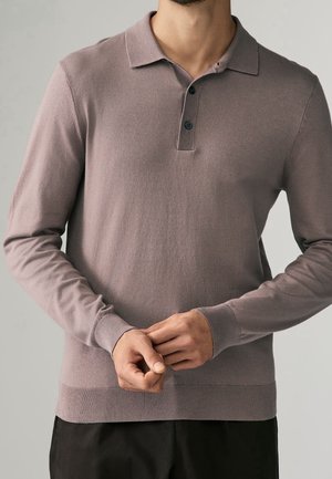 Mężczyzna ubrany w mauve polo z długim rękawem i kołnierzykiem zapinanym na guziki, z rękami swobodnie splecionymi na przodzie, na jednolitym tle.