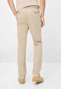 Beige Hosen mit geradem Schnitt, ausgestattet mit zwei hinteren Taschen mit Knöpfen und einer glatten Textur. Kombiniert mit hellbraunen Schuhen.