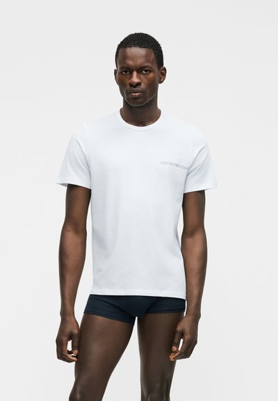 Moški stoji, oblečen v belo majico Emporio Armani in črne bokserice, na svetli enobarvni podlagi.