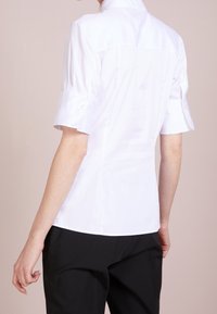 Camicetta bianca a maniche corte con colletto alto, tessuto liscio, dettagli a bottoni sulle maniche e una vestibilità sagomata sul retro.