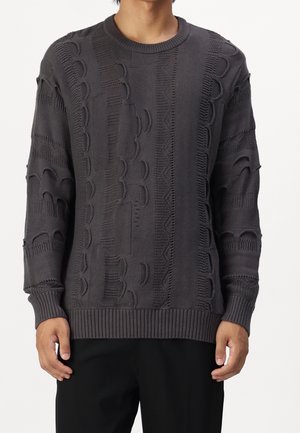 Homme portant un pull en maille gris foncé avec des motifs en relief texturés et des poignets et ourlet côtelés, associé à un pantalon noir.