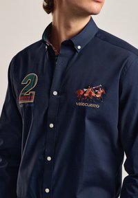 Camisa de botones azul marino con un cuello de cuadro contrastante, diseño bordado de carrera de caballos, número "2" y texto "valecuatro".