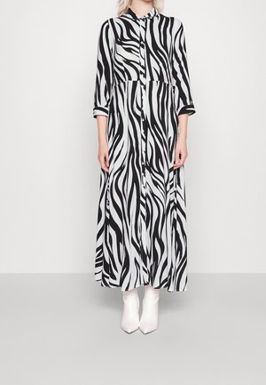 Personne portant une longue robe à imprimé zèbre noir et blanc avec des boutons et des bottines blanches, debout devant un fond uni.