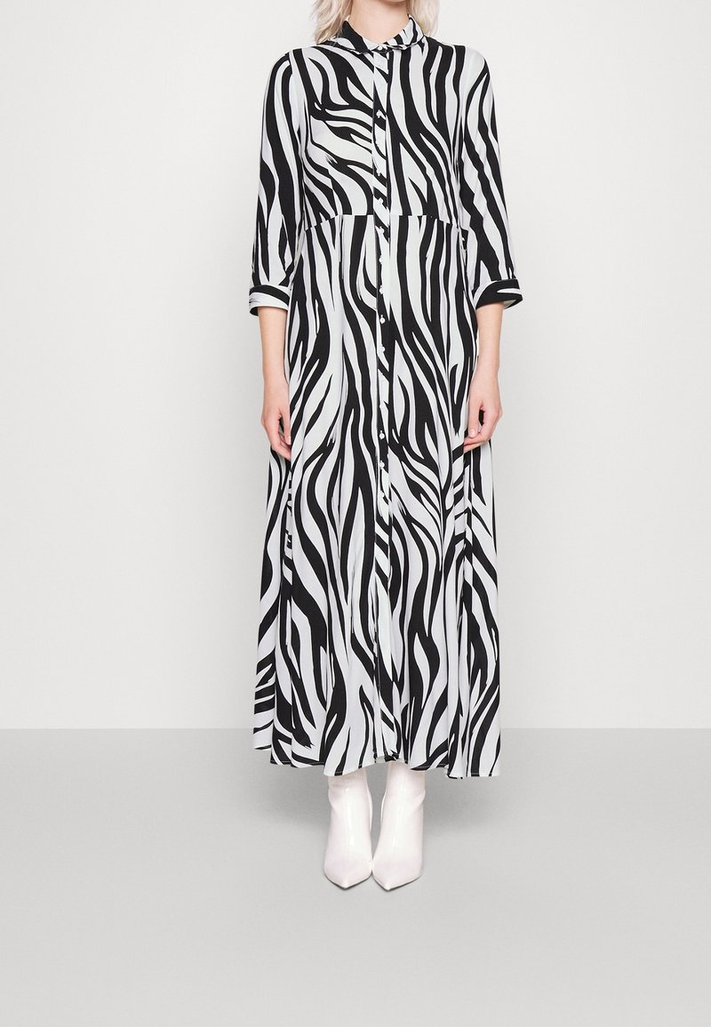 Personne portant une longue robe à imprimé zèbre noir et blanc avec des boutons et des bottines blanches, debout devant un fond uni.