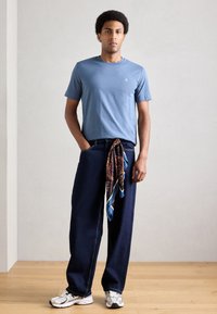 Gaiši zila īsroku T-krekls, tumši zili vaļīgi džinsi, pie jostas sasieta rakstaina šalle, balti sporta apavi, stāv uz koka grīdas pret neitrālu fonu.