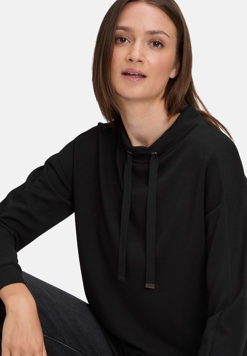 Czarna, długosleeve bluza z kapturem o wysokim dekolcie, z troczkami i metalowymi akcentami. Miękka tkanina i luźny krój, noszona na niebieskich dżinsach.