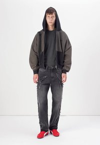 Sweatshirt oversized cinza-marrom com capuz preto, combinado com jeans cinza desgastados que apresentam detalhes em franjas. Ténis de desporto vermelhos completam o look.