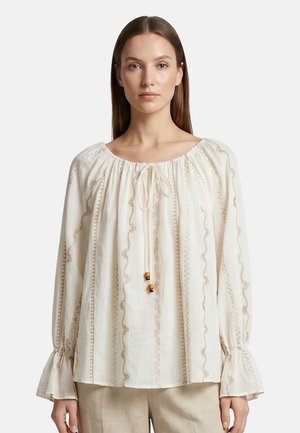 Vrouw met stijl bruin haar draagt een crèmekleurige blouse met beige borduurwerk, losse lange mouwen en een gestrikte halslijn met houten kralen.