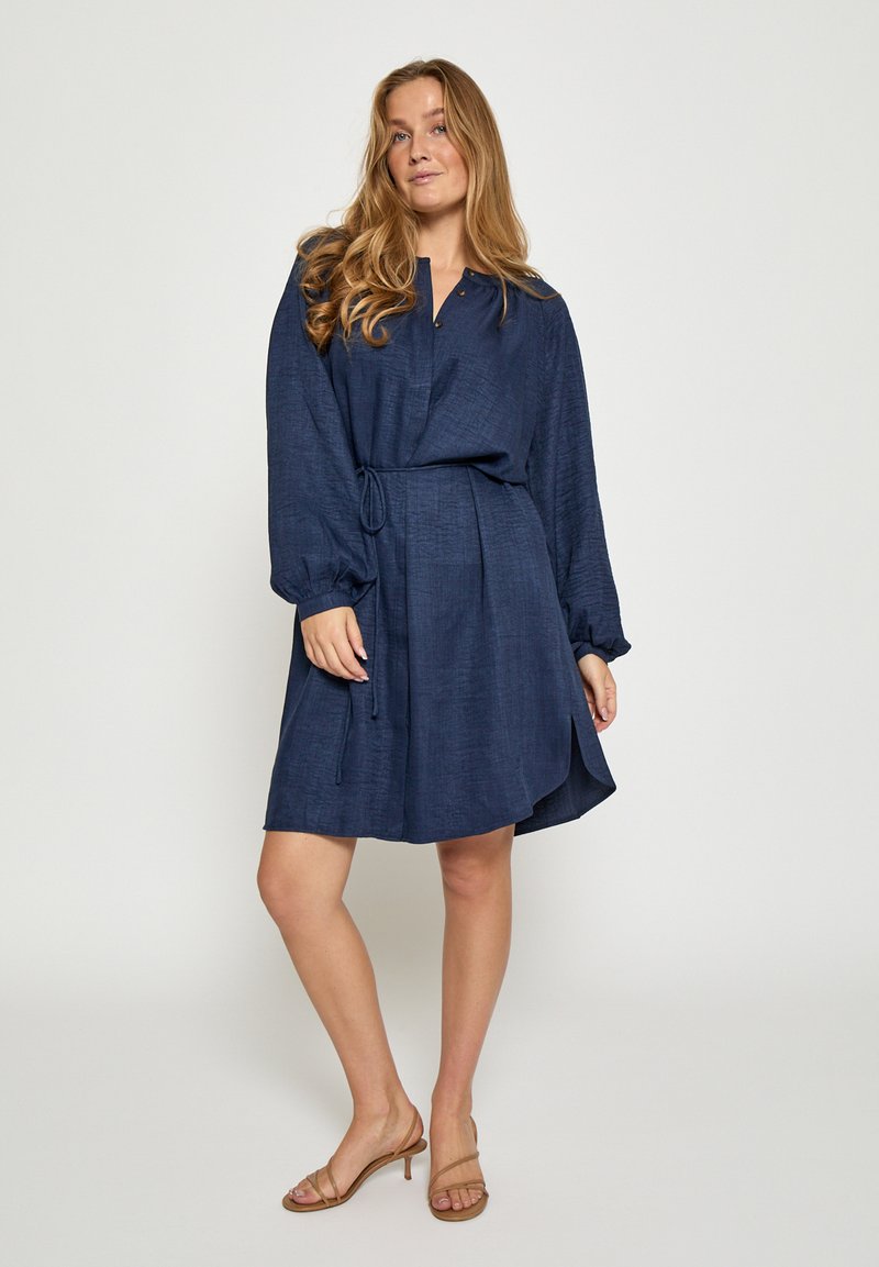 Temno modra obleka z ohlapnim krojem, dolgimi rokavi, ovratnikom z gumbi in pasom za zavezovanje. Tkanina ima teksturirano površino.