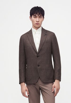 Junger Mann trägt einen braun karierten Blazer über einem weißen Hemd mit Knöpfen und beigen Hosen, steht vor einem schlichten weißen Hintergrund.