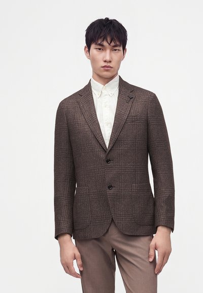 Jeune homme portant un blazer marron à carreaux sur une chemise blanche boutonnée et un pantalon beige, debout devant un fond blanc uni.