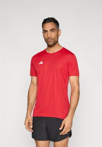 Punane lühikeste varrukatega sportlik särk, valmistatud kerge kangaga, millel on ümar kaelus ja valge kolme triibuga logo vasakul rinnal.