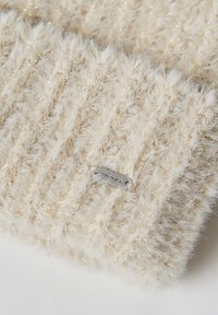 Cream-colored knitted beanie with a soft, fluffy texture and metallic gold threads. Features a metal brand tag attached to the fabric.