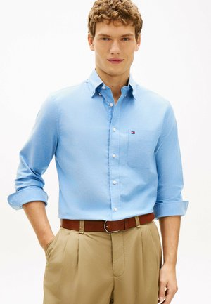 Młody mężczyzna ubrany w jasnoniebieską koszulę z logo na kieszeni i beżowe plisowane spodnie z brązowym paskiem, stoi z jedną ręką w kieszeni.