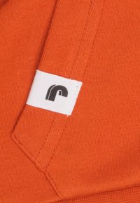 Oranje stof met een gladde textuur; heeft een wit label met een zwart, kleinletterig "r" logo; stiksels zijn zichtbaar langs de randen.