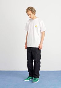 Fehér pamut póló, kis napgrafikával, fekete cargo nadrággal és zöld sportcipővel párosítva. Egyszerű, laza ruházat, látható minták nélkül.