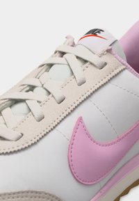 Λευκό και μπεζ αθλητικό παπούτσι με ροζ λογότυπο Nike, κρέμ κορδόνια και μαύρο και κόκκινο λογότυπο Nike στη γλώσσα.