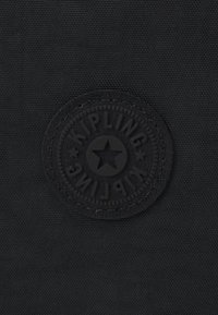 Black fabric patch featuring a round, embossed Kipling logo with a star in the center and circular border. Textured background.
