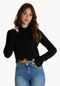 Maglione nero corto in tessuto strutturato con collo, quattro bottoni e orlo sfrangiato. Abbinato a jeans blu a vita alta.