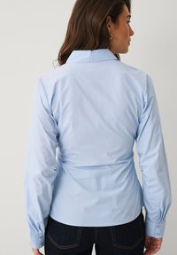 Chemise bleu clair à manches longues avec un col structuré, une taille cintrée et des poignets boutonnés, confectionnée dans un tissu lisse à texture minimale.