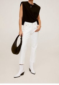 Top nero senza maniche, pantaloni bianchi a vita alta, stivaletti bianchi con punta a punta; tiene in mano una borsa nera curva.