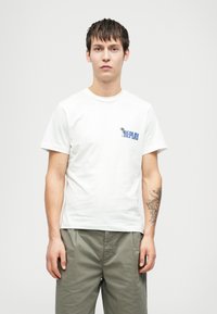 Jauns vīrietis ar īsiem tumšiem matiem, ģērbies baltā Replay T-kreklā un olīvzaļās biksēs, stāv pretī vienkāršai gaišai fona.