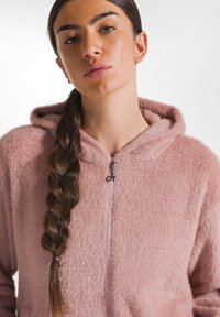 Roze fluffy zip-up hoodie met een zachte textuur, voorzien van een capuchon en een zilveren rits. Het ontwerp bevat een gevlochten haardetail.