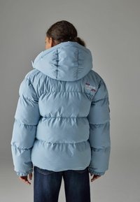 Doudoune bleu clair avec une grande capuche, design matelassé horizontal et poignets élastiques. Logo au dos en couleurs contrastées.