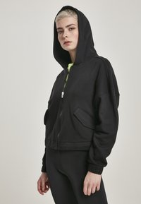 Hoodie nero cropped con zip frontale, grande cappuccio e due tasche laterali. Realizzato in morbido tessuto con polsini elastici e un inner layer verde neon.