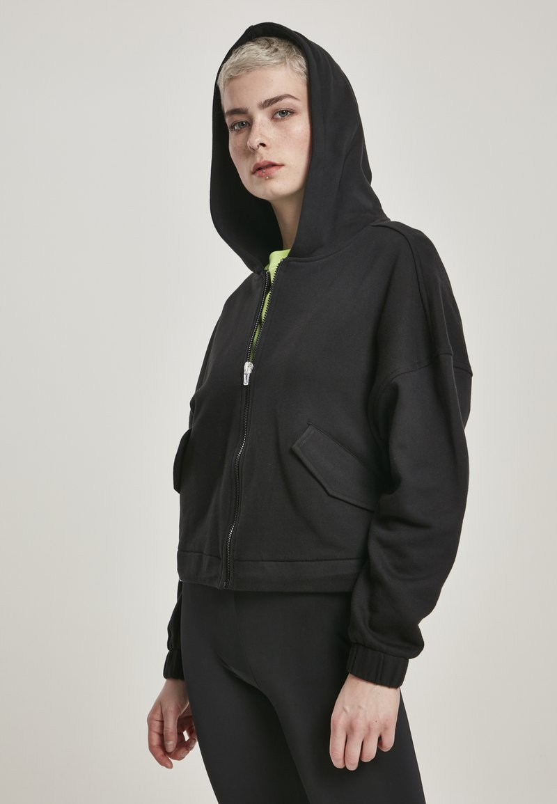 Hoodie nero cropped con zip frontale, grande cappuccio e due tasche laterali. Realizzato in morbido tessuto con polsini elastici e un inner layer verde neon.