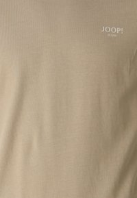 Beige kortærmet skjorte lavet af glat stof. Har et lille hvidt logo med teksten "JOOP! JEANS" på den øverste venstre side.