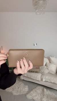 Portefeuille en cuir beige avec une fermeture éclair dorée et un logo embossé. Il présente une forme rectangulaire, une texture lisse et un design minimaliste.