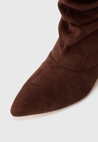 Barna velúr bokacsizma hegyes orral és ráncolt felső résszel, sima textúrával és varrott részletekkel az alján.
