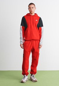 Bărbat tânăr în picioare, îmbrăcat cu un hanorac Nike roșu, negru și alb, pantaloni de trening roșii asortați și adidași albi, stând pe un podea verde.