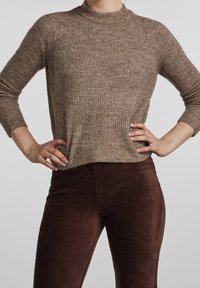 Brauner Rippstrickpullover mit hohem Kragen und 3/4-Ärmeln, kombiniert mit dunkelbraunen Cordhosen, die eine strukturierte Oberfläche aufweisen.