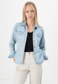 Camisa de mezclilla azul claro con dos bolsillos en el pecho, frente abotonado y mangas enrolladas, combinada con una camiseta negra y pantalones beige.