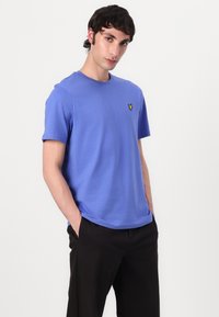 T-shirt en coton bleu clair à manches courtes, col rond et petit logo jaune brodé sur la poitrine. Porté avec un pantalon noir.