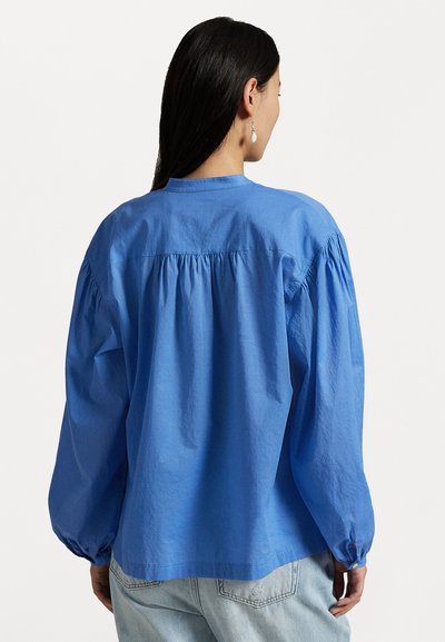 Blue cotton shirt with long puff sleeves, gathered yoke detail, and buttoned cuffs, paired with light-colored denim jeans.