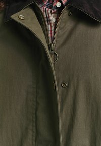 Unzipped olivgrüner Jacke mit bronzefarbenen Druckknöpfen, die den karierten Kragen eines Hemdes darunter zeigt, mit rot, weiß und schwarzen Karos.