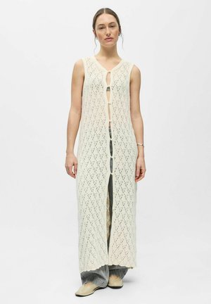 Kvinde iført en ærmeløs, lang cremefarvet strikket vest med diamantmønster, knaplukning foran, grå bukser og beige sko, stående mod hvid baggrund.