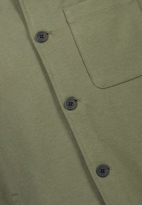 Camisa verde oliva con una textura suave, que incluye un bolsillo en el pecho y cuatro botones negros en la parte delantera.