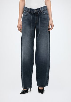 Femme portant un jean taille haute à jambes larges bleu foncé avec une ceinture nouée sur le devant et des talons noirs pointus sur un fond uni.