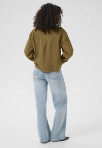 Olijfgroen, losvallend shirt met een achternaad en drop schouders, gecombineerd met lichtblauwe, wijde jeans en zwarte puntige schoenen.