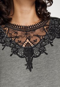 Camisa gris con un prominente escote de encaje negro con patrones florales y detalles de bordado intrincados que se extienden desde el cuello.