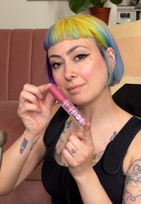 NYX This Is Milky Gloss dans un tube rose, avec une finition brillante, tenu par une personne avec des cheveux multicolores vibrants et des tatouages.