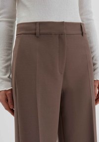 Braune maßgeschneiderte Hosen mit glatter Textur, gerade geschnitten, mit Gürtelschlaufen und seitlichen Taschen, kombiniert mit einem weißen strukturierten Oberteil.